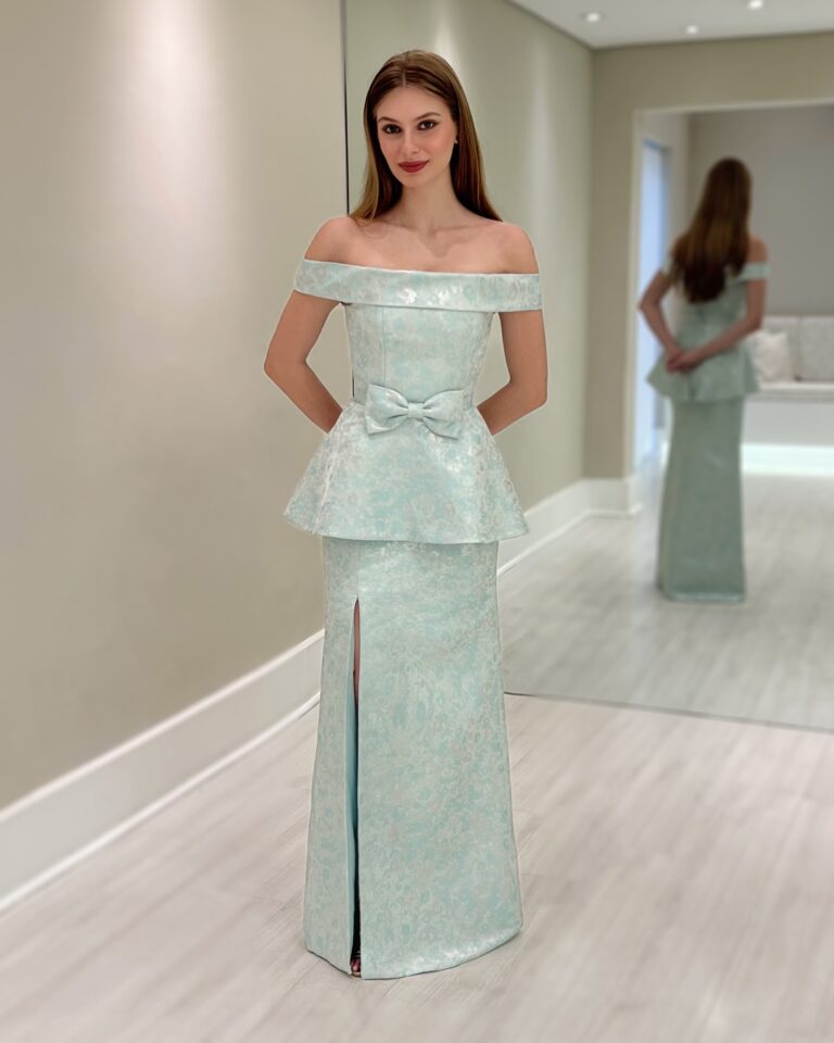 Modelo usando vestido longo verde em crepe com decote ombro a ombro, basque, laço na cintura e fenda frontal